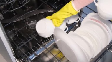 Bir kadın lastik eldiven verip, beyaz tabak ve bardakları bulaşık makinesine koyar. İleri teknolojileri kullanarak yüksek kaliteli mutfak gereçleri.
