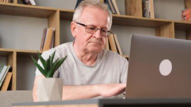 Gri saçlı, modern büyükbaba bilgisayarda yazı yazıyor ve yorgun, dizüstü bilgisayarı kapatıyor. Oturma odasındaki bilgisayarda çalışan yaşlı bir adam kitapların arka planına karşı çalışıyor. Dikey video
