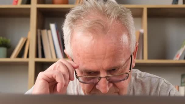 古い灰色の髪型の年金者は ラップトップモニターを見て疲れ メガネを外しました 高齢者は 本の背景に対してリビングルームのコンピュータで働いています バーティカルビデオ — ストック動画
