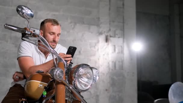 一个男人坐在车库的一辆复古摩托车上 手里拿着一部智能手机 漂亮的摩托车 — 图库视频影像
