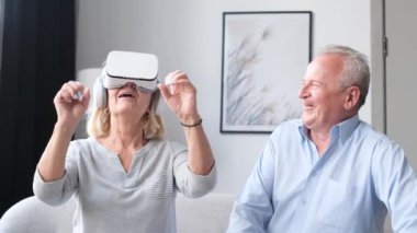 An elderly man happily looks at his wife wearing VR glasses. New technologies for entertainment in your home. Senior citizen wearing VR headset or visual reality glasses.
