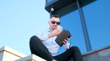 A young man is stylishly dressed and wearing safety glasses counts money against the background of a business center. Concept of finance