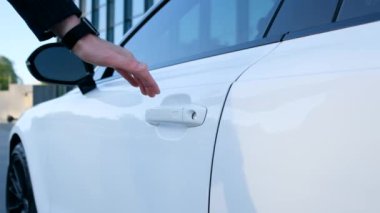 A close-up of a man opens the door of a white car. The driver gets behind the wheel.