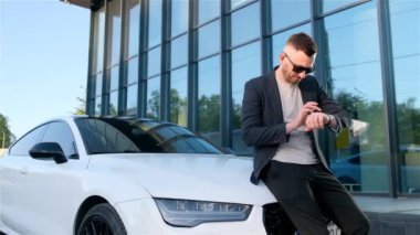 a businessman looks at a smart watch while standing near his car.