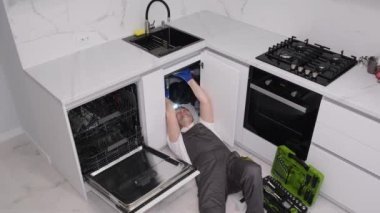 A handyman fixes a water leak from under the sink. A plumber lies under a sink to repair a pipe.