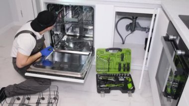 A young repairman is repairing a broken dishwasher in a modern kitchen.