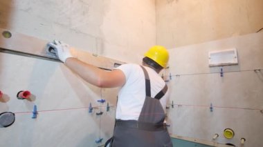 A builder in overalls is installing ceramic tiles in a bathroom. Repair in a new building.