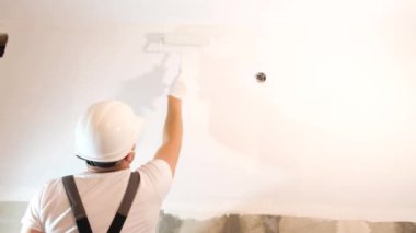 A man paints the wall white with a roller. Concept builder or artist in helmet with paint roller over empty room. 4k video