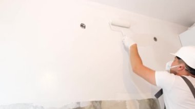 A young man in a construction helmet paints the walls in an apartment.