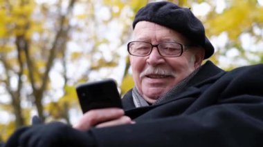 Portrait of an old man in glasses on the background of an autumn tree, sitting and looking at a smartphone. Elderly people and technology