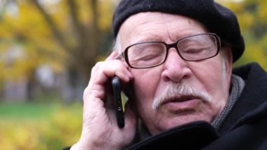 A pleasant old gray-haired man in glasses is sitting on a park bench and talking on the phone. Autumn walk in the park.