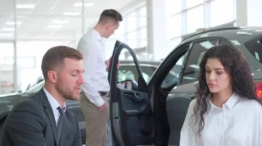 A car dealer consults a young couple in a car dealership. Professional employee helping with the selection of a new car, talking and gesturing. 4k video