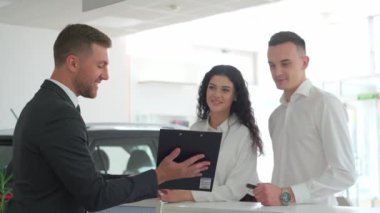 A young couple signs an order for the purchase of a car at the companys office. A car dealer works with a European couple at a car dealership and talks about buying a new car. Salesman.