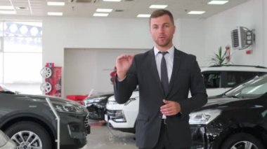 A young man poses for new cars in a car showroom. He is standing between new cars, looking at the camera and talking. Sale of modern cars