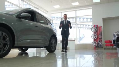 A young man poses for new cars in a car showroom. He is standing between new cars, looking at the camera and talking. Sale of modern cars