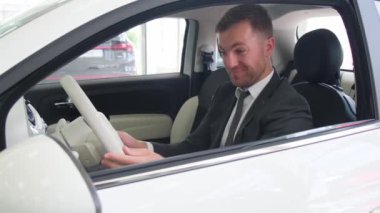 European man on a new car in a car showroom, portrait of a man in a car. He is satisfied with the purchase of a car