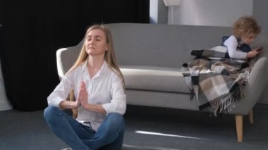 The young mother is doing yoga and the child is sitting on the sofa. Relaxation at home.