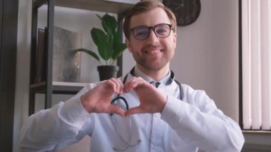 Modern ofisinde kameraya gülümseyen Avrupalı bir doktorun portresi. Kardiyolog kendi elleriyle kalp hareketi gösterdi.