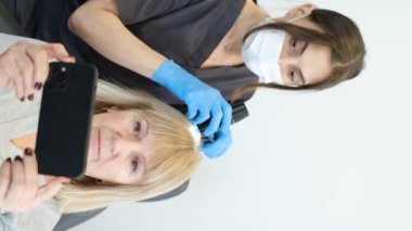 Genç bir trikoloğun yaşlı bir hastanın kafasını inceleyen ve telefonda yayın yapan dikey videosu. Saç ve kafa derisine zamanında müdahale.