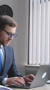 Handsome manager working with laptop in office. Confident businessman sitting at the office table and working with a laptop, he is typing. Vertical video