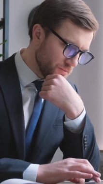  Handsome bearded manager wearing glasses sitting in office using his laptop and working with computer program. Vertical video