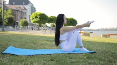 Sportif kadın egzersiz minderinde bacaklarını esnetiyor, olumlu gülümsüyor, esneklik egzersizleri yapıyor, temiz havanın tadını çıkarıyor.