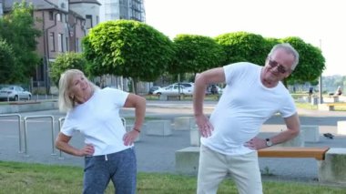 Gülümseyen mutlu aile çifti spor kıyafetleri içinde güneşli bir sabahta parkta birlikte egzersiz yaparken kollarını esnetiyorlar..
