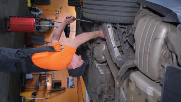 車の修理工場で働く制服の若いメカニックの垂直ビデオを持ち上げられた車 — ストック動画