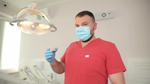 歯科医は歯科インプラントを手に持っていて カメラを見ています 赤い医療コートの男性歯科医の肖像画 — ストック動画