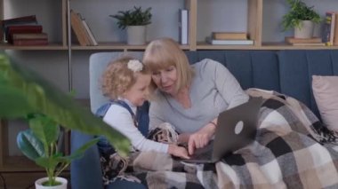 A grandmother spends time with her granddaughter in the living room, they use a laptop. Happy family values. They laugh merrily