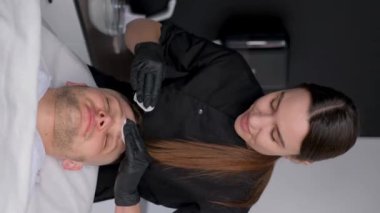 Dikey video. Güzellik uzmanı, kuaförde pamuklu süngerle müşterinin suratını siliyor. Dermatologda genç bir adam. Yüz derisi için tedavi prosedürleri