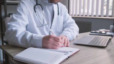 Gözlüklü ve üniformalı profesyonel genç doktor ofisinde evrak işleriyle uğraşıyor. Teşhis, tedavi planı yazıyorum. İş yerinde bir doktor. Dikey video