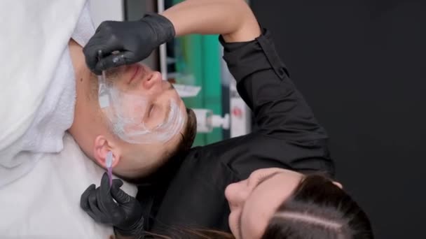 一个戴手套的美容师把面具戴在男人脸上的垂直录像 男性面部皮肤护理 治疗口罩 — 图库视频影像
