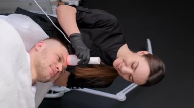 Dikey video. Elektronik çok fonksiyonlu masaj aleti kullanarak erkek yüz masajı. Kozmetik uzmanı bir kız genç ve yakışıklı bir adam için gençleştirici prosedürler uyguluyor.