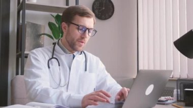 Avrupa görünümlü genç bir doktor ofiste oturuyor ve bilgisayar başında çalışıyor. Doktor dizüstü bilgisayardaki işini bitirir. Yoğun bir iş. Yorgun doktor. Baş ağrısı. Dikey video
