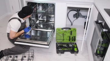 A young repairman is repairing a broken dishwasher in a modern kitchen.