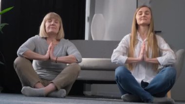 An elderly mother does yoga with her daughter. Active woman with daughter practicing lotus pose while sitting on yoga mat at home.