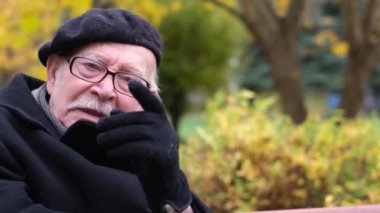 Close-up portrait of an old man in a beret against the background of an autumn tree, sitting on a bench and talking to the camera. Pensioners. Seniors