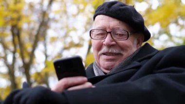 Portrait of an old man in glasses on the background of an autumn tree, sitting and looking at a smartphone. Elderly people and technology