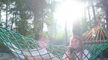 Young beautiful woman laughs while talking on the phone while lying in a hammock in a pine forest. Rest in the village during the summer holidays.