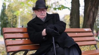 An elderly man in black clothes sits on a park bench and puts on black gloves. Rest in the autumn park.