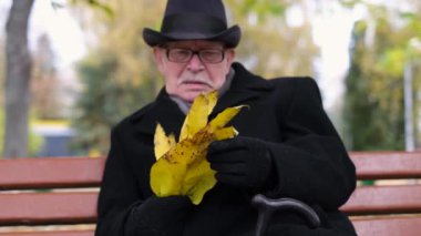 An old lonely man in a black coat and hat sits in an autumn park and holds yellow leaves. Happy elderly people