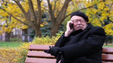 A pleasant old gray-haired man in glasses is sitting on a park bench and talking on the phone. Autumn walk in the park.
