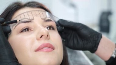 A close-up beautician corrects the shape of the eyebrows with a special ruler. Beautiful girl in a beauty salon