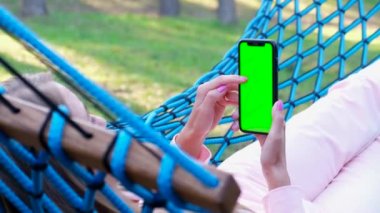 A young woman is resting in the forest, she is lying in a hammock and using a smartphone with a green screen. Outdoor recreation. Chromakey background