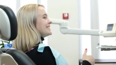 Dişçi koltuğunda güzel beyaz gülüşlü, baş parmağını gösteren genç sarışın kadın. Dişçinin memnun müşterisi.