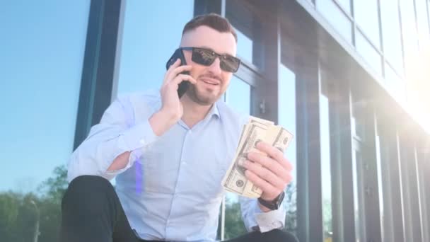 彼の手にドルを持った若いボスがスマートフォンで話している ビジネスマネージャーのコンセプト バーティカルビデオ — ストック動画