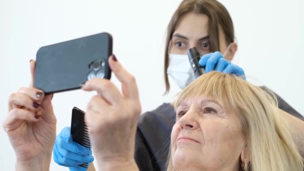 一位老年妇女的头皮由办公室里的一位三位一体的医生治疗 一位女性三叶草学家给病人剃头 并在手机上播放视频 垂直录像 — 图库视频影像