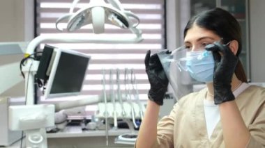 Genç çekici esmer dişçi, dişçi muayenesinden önce koruyucu gözlük takıyor ve iş yerinde kameraya bakıyor. Diş hekimliğinde modern yaklaşımlar