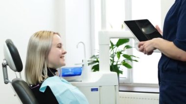 Modern bir ofiste dişçi randevusunda pozitif sarışın. Kadın bir dişçi, tablet kullanan bir hastaya danışır..